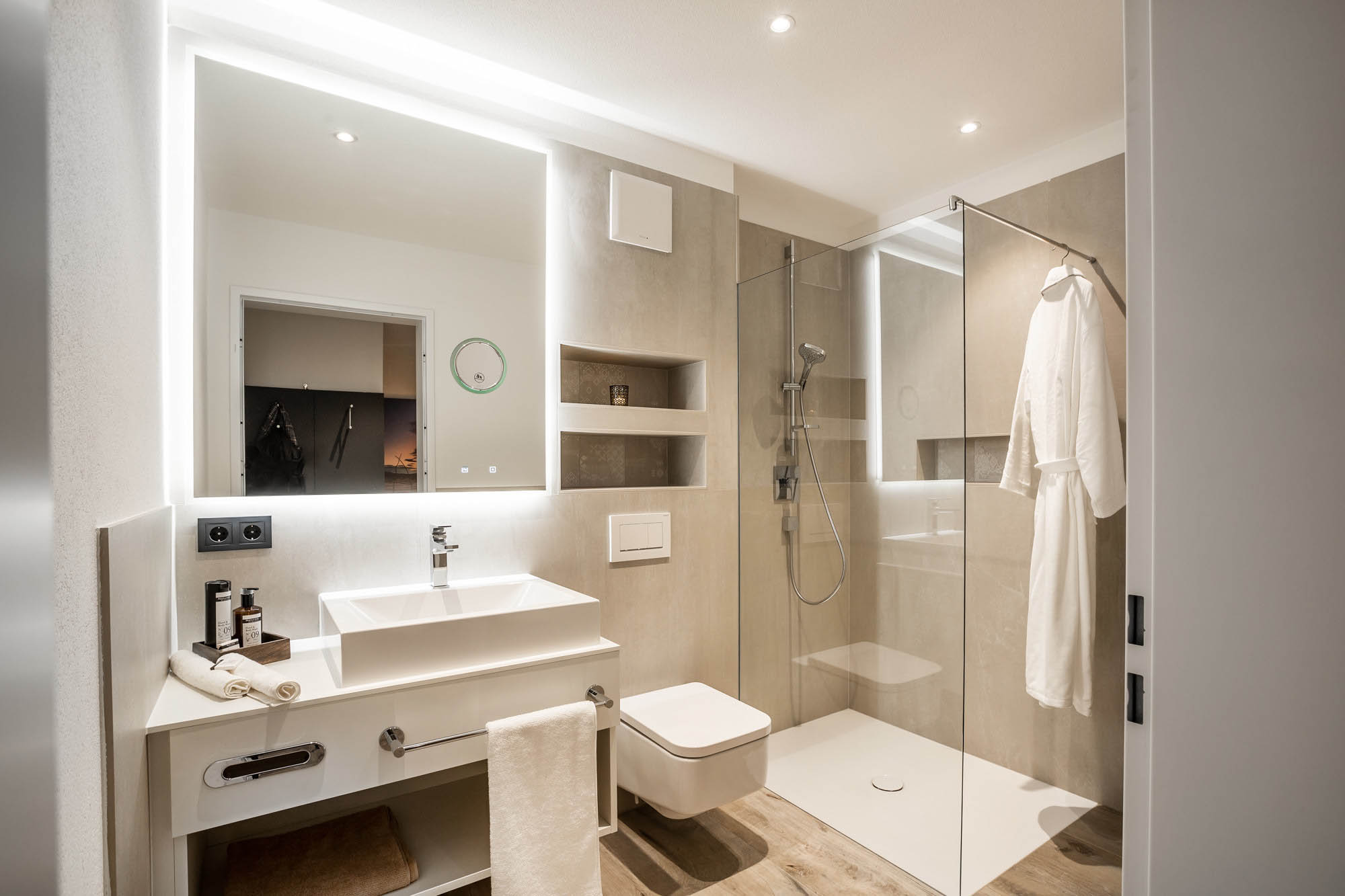 Blick ins Comfortbad mit ebenerdiger Dusche mit Glastrennwand, einer Toilette und einem Waschtisch mit weißem Waschbecken und großem Spiegel. 